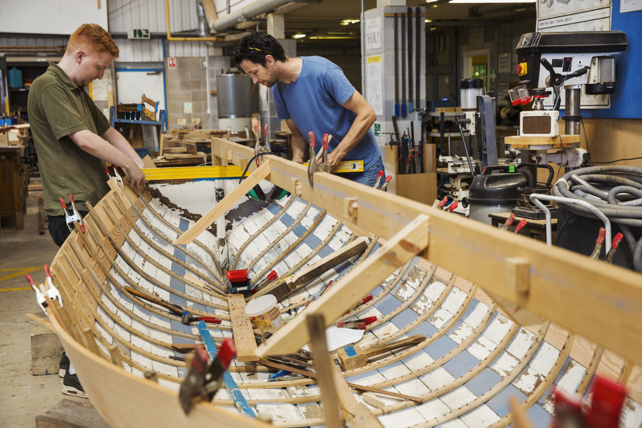 Two men in a boat-builder's workshop, working together on a wooden boat hull.